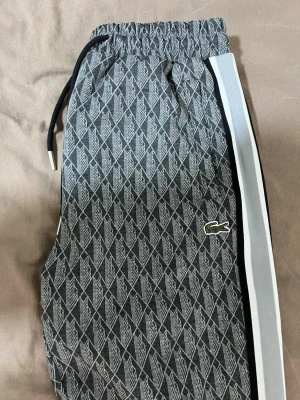 Grå mönstrade mjukisbyxor Lacoste - Säljer ett par grå mjukisbyxor från Lacoste med vitt och svart sidoband och ett diskret mönster över hela byxan. Byxorna har dragsko i midjan och den klassiska Lacoste-loggan på benet. Perfekta inför sommaren samtidigt som en chill och trendig look.