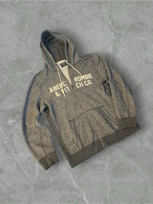 Grå hoodie från Abercrombie & Fitch | M - Snygg grå zip-hoodie från Abercrombie & Fitch med vita dragsnören och tryckt logga på bröstet. Hoodien har två fickor framtill och ribbade muddar vid ärmar och nederkant. Perfekt för en avslappnad och trendig look.
