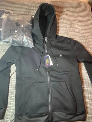Svart hoodie från Polo Ralph Lauren - Svart hoodie med dragkedja från Polo Ralph Lauren. Klassisk design med huva och dragsnören, två fickor framtill och den ikoniska lilla loggan broderad på bröstet. Perfekt för en chill och stilren look. Passar S oh M!