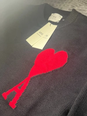 Svart stickad tröja från Ami Paris - Svart stickad tröja från Ami Paris med deras ikoniska röda hjärta och A-logga på bröstet. Tröjan har rund halsringning, ribbade muddar och är tillverkad i mjukt material. Perfekt för dig som gillar stilrena och trendiga plagg.