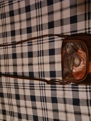 Brun axelväska i läder med patchwork - Snygg axelväska i brunt och mörkbrunt läder med patchwork-design och justerbar rem. Väskan har ett dekorativt metallspänne framtill och flera fack. Perfekt för dig som gillar retrostil och unika detaljer.