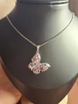Silverhalsband med lila fjärilshänge - Ett elegant halsband i silverfärg med ett fjärilsformat hänge dekorerat med lila stenar och små glittrande detaljer. Perfekt för dig som gillar unika smycken med lite extra bling och en feminin touch. Kedjan är tunn och smidig, och hänget har en snygg form.