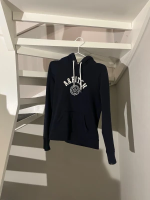 Mörk blå hoodie från Abercrombie & Fitch - Väldigt trendig hoodie från Abercrombie & Fitch med vit logga. Den passar som XS även fast det står L i hoodien. Perfelt för en avslappnad och trendig stil. Inga defekter och priset är inte hugget i sten vid en smidig affär!