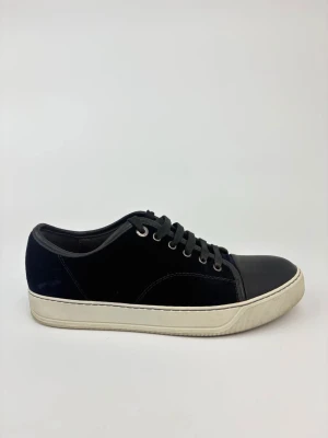 Svarta Lanvin sneakers med vit sula - Stilrena svarta sneakers med vit sula och svarta skosnören. Ovandelen är i mockaliknande material och tån är förstärkt med slätare svart material. Klassisk låg modell med metallhål för snörning och diskret design.