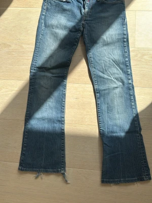 Blå bootcut jeans med slitna benslut - Ett par blå bootcut jeans med slitna och fransiga benslut. Jeansen har klassisk femficksdesign och dekorativa sömmar på bakfickorna. Perfekta för en avslappnad och trendig look.