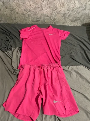 Nike miller shorts o tshirt - Säljer ett snyggt rosa träningsset från Nike med t-shirt och shorts. Båda plaggen har Nike-logga i silver och är tillverkade i ett lätt och snabbtorkande material. Perfekt för träning eller sportaktiviteter. T-shirten har rund hals och shortsen har resår i midjan med dragsko.