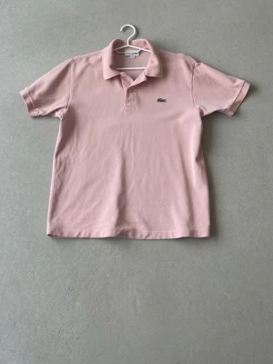 Rosa pikétröja från Lacoste - Klassisk ljusrosa pikétröja från Lacoste med korta ärmar och krage. Tröjan har den ikoniska krokodilloggan på bröstet och är tillverkad i mjuk bomull. Perfekt för en avslappnad och stilren look.