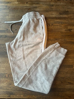 Grå mjukisbyxor från Adidas - Gråa mjukisbyxor från Adidas med vita ränder längs sidorna och broderad logga på benet. Byxorna har resår och dragsko i midjan samt muddar vid bensluten. Tillverkade i mjukt och bekvämt material, perfekta för en avslappnad stil.