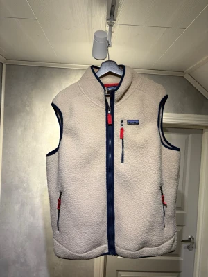 Beige fleeceväst från Patagonia - Säljer en beige fleeceväst från Patagonia med marinblå detaljer och röda dragkedjor. Västen har hel dragkedja framtill, två sidofickor och en bröstficka. Perfekt för lager-på-lager och outdoor-stil. Helt ny aldrig använd, storlek XL. Längd 71cm, axel, 46cm, bröst 61cm skickas samma dag eller dagen efter köp