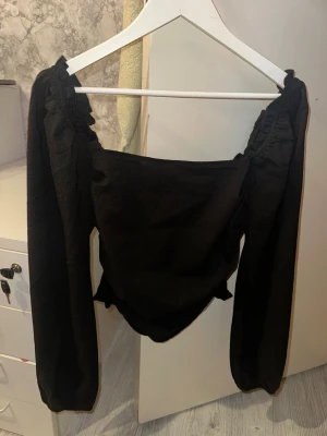 Svart offshoulder topp med långa ärmar - Trendig svart offshoulder topp med långa puffärmar och smockad rygg. Toppen har en croppad passform och är tillverkad i ett mjukt, lätt material som ger en snygg siluett. 