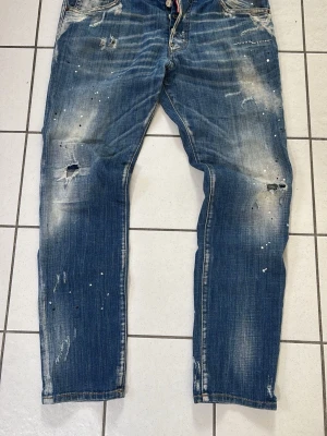 Dsquared2 jeans  - Säljer ett par blå jeans från Dsquared2 med slitna detaljer, hål och färgstänk för en cool, trendig look. Jeansen har raka ben, klassisk femficksmodell och Dsquared2-logga bak. Perfekta för dig som gillar streetstyle och unika plagg.