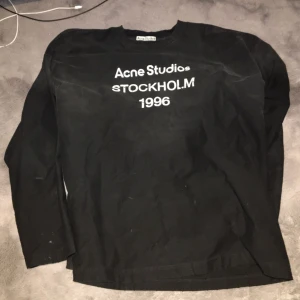 Svart långärmad topp Acne Studios - Svart långärmad topp från Acne Studios med vit text 'Acne Studios STOCKHOLM 1996' på bröstet. Klassisk rund halsringning och rak passform. Perfekt för en stilren och trendig look.