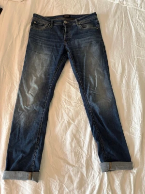 Mörkblå jeans Jack&Jones - Snygga mörkblå jeans med klassisk femficksdesign och uppvikta benslut. Jeansen har en lätt tvättad look framtill och raka ben. Perfekta för en avslappnad stil och passar till det mesta. Materialet är denim i bomull. Storlek 32-34