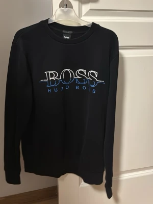 Svart sweatshirt från Hugo Boss - Svart sweatshirt med rund hals från Hugo Boss. Framsidan har en stor vit och blå BOSS-logga samt texten HUGO BOSS i blått. Tröjan har långa ärmar och ribbade muddar vid ärmslut och nederkant. Perfekt för en stilren och avslappnad look.