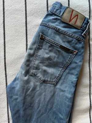 Blå jeans från Nudie Jeans - Säljer ett par klassiska blå jeans från Nudie Jeans med snygg tvätt och diskreta slitningar. Jeansen har fem fickor, orangea sömmar och en patch med logga bak i midjan. Tillverkade i bomull och har en rak passform som funkar till det mesta.