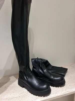 Svarta overknee chunky stövlar stl 39 - Säljer ett par svarta overknee stövlar med chunky sula och dragkedja på insidan. Stövlarna är i slätt konstläder och har en modern, hög passform som går över knät. Perfekta för att lyfta vilken outfit som helst. Använda en gång helt nya endast en liten repa (se tredje bilden) nypris 699:-