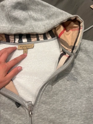 Grå Burberry hoodie med dragkedja "ÄKTA" PRISET DISKUTERAS💯 - Snygg grå hoodie från Burberry med dragkedja och klassiskt rutigt foder i huvan. Mjuk insida i fleece och broderad logga på bröstet. Perfekt för dig som vill ha en stilren och lyxig look med ikoniska Burberry-detaljer.