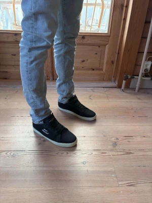 Svarta Saint Laurent sneakers - Svarta sneakers från Saint Laurent med vit sula och tydlig logga på sidan. Skorna har klassisk låg modell, svarta skosnören och detaljer i guld på plösen. Materialet är canvas och gummi. Perfekta för en stilren och trendig look. Skorna har tydliga slitningar, menade såklart. Skorna är i nyskick (9,5/10). Dustbag samt köparkort medföljer.