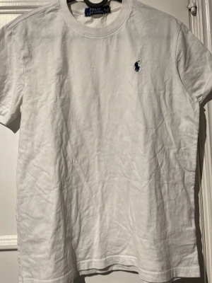 Vit t-shirt från Polo Ralph Lauren - Klassisk vit t-shirt från Polo Ralph Lauren med rund halsringning och korta ärmar. T-shirten har en broderad marinblå logga på bröstet och är tillverkad i mjuk bomull. Perfekt basplagg med stilren design.