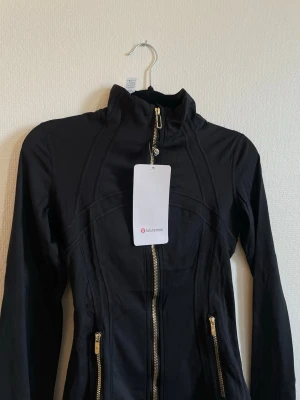 Svart träningsjacka från Lululemon st 4 - Snygg svart träningsjacka från Lululemon med guldiga dragkedjor och hög krage. Jackan har figurnära passform, långa ärmar och två fickor framtill. Perfekt för gymmet eller löprundan. Materialet är stretchigt och följsamt.