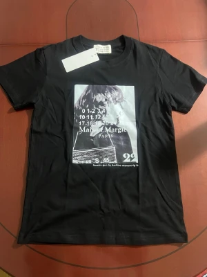 Maison Margiela svart t-shirt med print - Svart t-shirt från Maison Margiela med ett stort grafiskt print framtill i svartvitt. Klassisk rund halsringning och korta ärmar. T-shirten har en modern och edgy vibe, perfekt för dig som gillar statement-plagg och streetstyle. OBS! Tröjan är lite tightare runt bröstet och kan därmed passa även som S