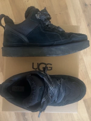  UGGs lowmels  - Säljer ett par svarta skor från UGG med robust sula och snygga detaljer. Ovandelen är i mocka och textil, med vadderad insida för extra komfort. Skorna har grova snören och metallhakar för en cool look. Perfekta för dig som vill ha både stil och funktion.