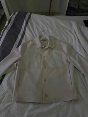 Beige overshirt från Zara - Stilren beige overshirt från Zara med raka linjer och två stora bröstfickor. Klassisk krage och knäppning framtill med matchande knappar. Perfekt för lager-på-lager och enkel att styla till olika outfits. Tillverkad i ett mjukt tyg som känns skönt mot huden.
