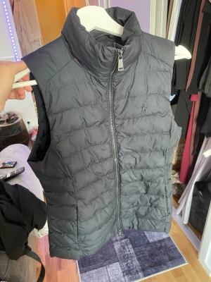 Svart dunväst från Polo Ralph Lauren - Snygg svart dunväst från Polo Ralph Lauren med hög krage och dragkedja framtill. Västen har en quiltad design och är tillverkad i ett lätt och värmande material, perfekt för lager på lager under kyliga dagar.