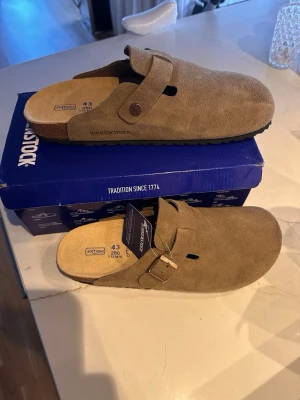 Birkenstock Boston tofflor mocka brun Helt nya - Säljer ett par Birkenstock Boston tofflor i brun mocka med justerbart spänne och mjuk fotbädd. Klassisk slip-in modell med svart räfflad gummisula för bra grepp. Perfekta för chill dagar hemma eller när du vill ha en avslappnad stil.
