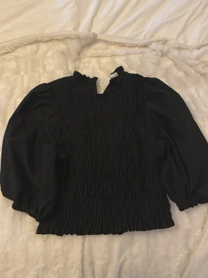 Svart blus med puffärmar och rynk - Trendig svart blus med rynkad framsida och volangdetaljer vid halsringningen. Blusen har långa puffärmar och elastisk nederkant som ger en snygg siluett. Perfekt att styla med jeans eller kjol för en cool look.