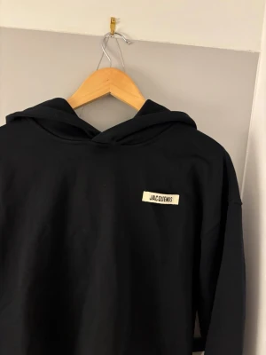 Svart hoodie från Jacquemus - Snygg svart hoodie från Jacquemus med minimalistisk design och liten vit logotyp på bröstet. Hoodien har huva och långa ärmar, perfekt för en clean och stilren look. Tillverkad i mjukt material som känns skönt mot huden. Priset är självklart diskuterbart