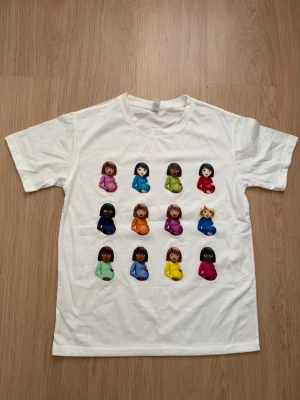 Vit t-shirt med färgglada emoji-tryck - Vit t-shirt med tryck av flera färgglada gravid-emoji i olika färger på framsidan. T-shirten har rund hals och korta ärmar, perfekt för dig som gillar unika och lekfulla plagg. Materialet känns mjukt och bekvämt. DRAKE