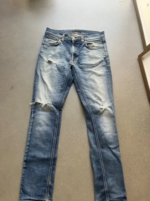 Ljus Blå Nudie jeans  - Säljer ett par blå jeans med slitningar och hål på knäna. Jeansen har en klassisk femficksmodell, raka ben och snyggt tvättad denim med ljusa partier. Jeansen är i storleken w30 l32. Hör av er vid minsta fundering om jeansen! MVH Elliot.