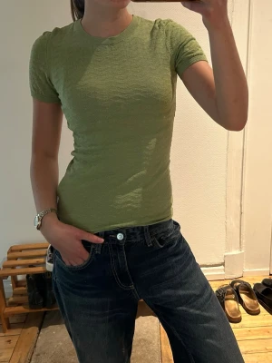 Ljusgrön tight t-shirt - En ljusgrön, tight t-shirt med korta ärmar och rund halsringning. T-shirten har en enkel och stilren design, perfekt att matcha med jeans eller kjol. Materialet ser ut att vara mjukt och stretchigt, vilket ger en bekväm passform.