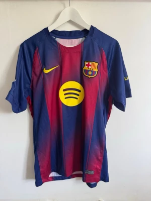 FC Barcelona matchtröja Nike - Säljer en FC Barcelona matchtröja i blått och rött med gula detaljer. Tröjan har klubbmärke, Nike-logga och Spotify som sponsor framtill. På ryggen står det Lamine Yamal och nummer 10 i gult. Materialet är lätt och andas, perfekt för fotboll.