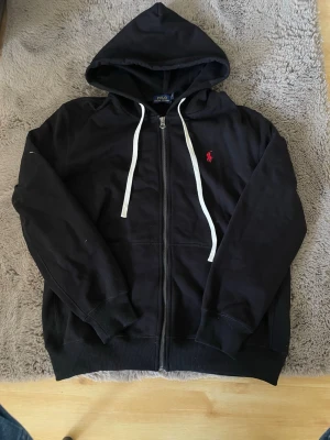 Svart hoodie från Polo Ralph Lauren - Snygg svart hoodie från Polo Ralph Lauren med dragkedja, vit dragsko och klassisk röd logga på bröstet. Tillverkad i mjuk bomullsmix och har en stor huva. Perfekt för en avslappnad och stilren look. Säljer för den ej passa mig 