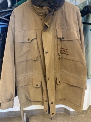 Beige parkajacka från Fjällräven - Klassisk beige parkajacka från Fjällräven med hög krage, flera stora fickor och broderad logga på bröstet. Jackan har långa ärmar, knappar och dragkedja framtill samt justerbar mudd vid ärmslut. Perfekt för dig som gillar outdoor och funktionell stil.