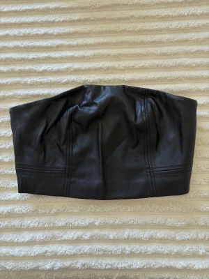 Svart skinn bandeau tubtopp - Svart bandeau tubtopp i skinnimitation med markerade sömmar och dragkedja bak. Enkel och stilren design utan axelband.