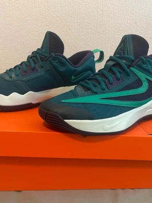 Nike gröna sneakers Giannis Immortality - Snygga sneakers från Nike Giannis Immortality-serien i mörkgrönt och turkost med vita detaljer. Skorna har mesh- och syntetmaterial för bra andningsförmåga och komfort. Låg modell med klassisk Nike Swoosh på sidan och Giannis-logga på plösen. Perfekta för en sportig look.