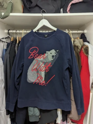 Mörkblå sweatshirt H&M med isbjörn - Mörkblå sweatshirt från H&M med ett stort tryck av en isbjörn och texten 'Bear With Me!' i rött framtill. Tröjan har rund halsringning och långa ärmar. Tillverkad i mjukt bomullsmaterial, perfekt för en avslappnad stil.