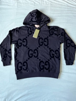 Svart hoodie från Gucci med GG-mönster - Snygg svart hoodie från Gucci med stort GG-mönster över hela tröjan. Klassisk huva och ribbade muddar vid ärmslut och nederkant. Perfekt för dig som vill sticka ut med en exklusiv streetstyle-look.