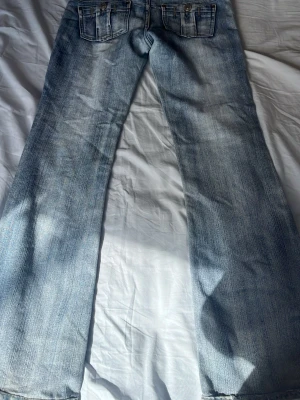 Ljusblå bootcut jeans med fickor - Säljer ett par ljusblå bootcut jeans med slitningar och två bakfickor med lock och knapp. Jeansen har klassisk femficksdesign och är tillverkade i denim med snygg tvättad look. Perfekta för dig som gillar retrostil och bredare ben.