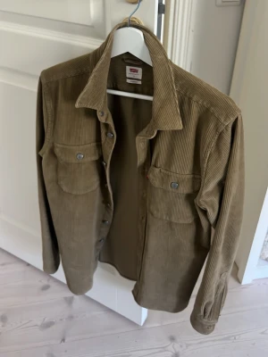 Beige manchester overshirt från Levi's - Snygg beige overshirt från Levi's i mjuk manchester. Skjortan har två bröstfickor med knappar, klassisk krage och raka ärmar. Perfekt lager-på-lager-plagg med avslappnad passform och cool vintagekänsla.