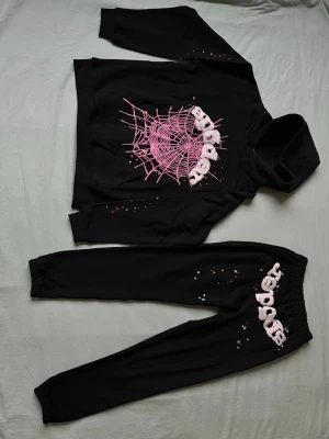 Svart loungewear-set Spider - Svart loungewear-set med hoodie och byxor. Båda plaggen har tryck med rosa spindelnät och texten 'spider' i vit fluffig stil. Färgglada små stjärnor och prickar som detaljer. Hoodien har magficka och byxorna resår i midjan.