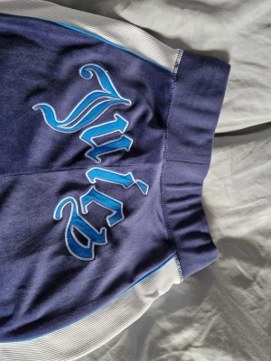 Navy blue velourbyxor från Juicy Couture - Mörkblå mjukisbyxor i velour från Juicy Couture med ljusblå sidor och stort 'Juicy'-tryck bak i blått och vitt. Byxorna har bred resår i midjan och raka ben. Perfekta för en avslappnad och trendig look. Limited edition i begränsat lager, storlek XS.