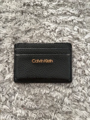 Svart korthållare från Calvin Klein - Snygg svart korthållare i präglat läder från Calvin Klein med guldfärgad logga framtill. Smidig och stilren design med flera kortfack både fram och bak. Perfekt för dig som vill ha ett minimalistiskt och trendigt accessoar.