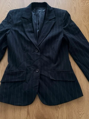 Svart kavaj med vita ränder - Snygg svart kavaj med tunna vita kritstrecksränder och klassisk krage. Två knappar framtill och fickor med lock. Perfekt för en stilren look. Materialet känns mjukt och följsamt.