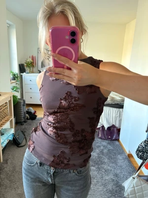 Brun blommig ärmlös topp - Säljer en brun ärmlös topp med stora blommiga broderier i mörkare nyans. Toppen har en tight passform och är tillverkad i ett stretchigt material som sitter snyggt på kroppen. Perfekt för dig som vill ha något unikt och trendigt i garderoben.