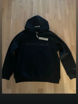 Svart Essentials hoodie med tryck - Svart hoodie från Essentials med stor logga tryckt på bröstet och etikett i nacken. Klassisk känguruficka framtill och huva. Perfekt för en avslappnad streetwear-look. Mjuk bomullsblandning för extra komfort.