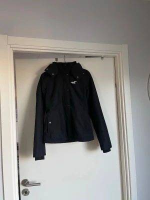 Svart vindjacka från Hollister - Snygg svart allväderjacka  från Hollister med huva och dragkedja samt knappar. Jackan har turkosa detaljer, inklusive loggan på bröstet, och praktiska fickor med knappar. Insidan är fodrad med mjukt fleece material för extra komfort.  Luvan är även fodrad Perfekt för blåsiga & regniga dagar. Perfekt vår och höst jacka 